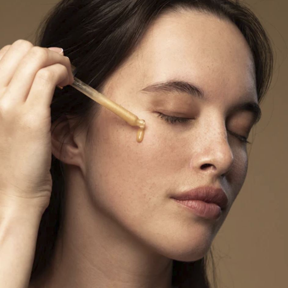 Woman applying a dropper of liquid to her face against a neutral background