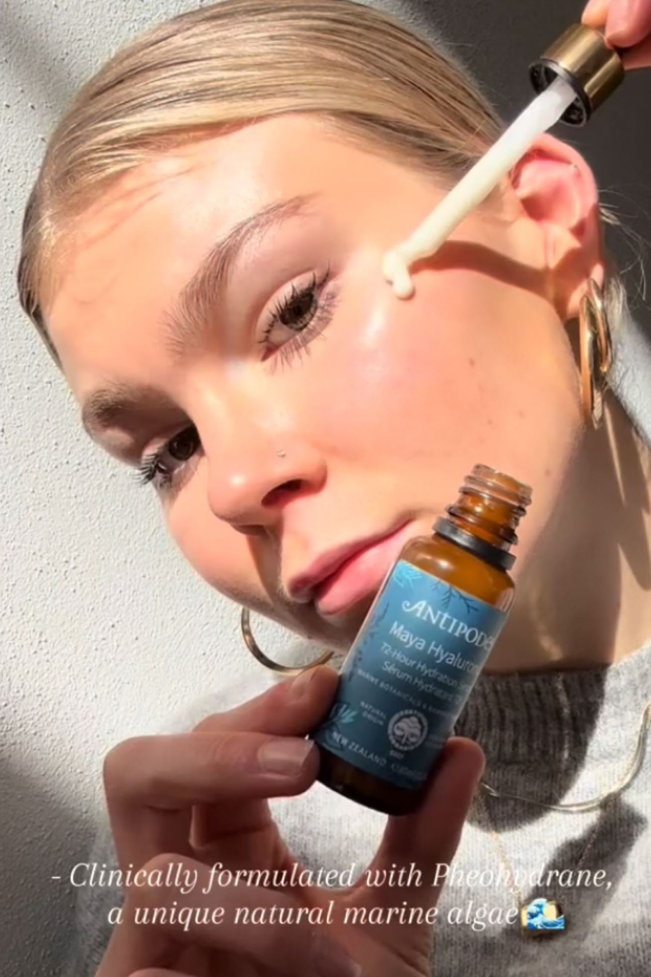 Person applying a dropper of skincare product to their face with Antipodes bottle in hand.
